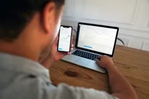 Young investor reviewing stock charts on phone and laptop, learning how to invest in their 20s